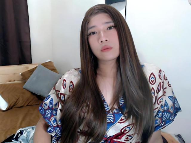 SophiaNicolex with long brown hair, wearing a patterned top, sitting on a bed with brown pillows, looking at the camera with a neutral expression.