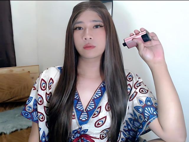 SophiaNicolex with long brown hair, wearing a colorful patterned dress, holding a pink and black vape pen, looking directly at the camera with a neutral expression.
