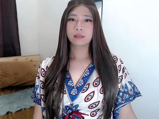 SophiaNicolex has long brown hair, fair skin, and is wearing a patterned dress with blue, red, and white peacock designs, showing a subtle smile.