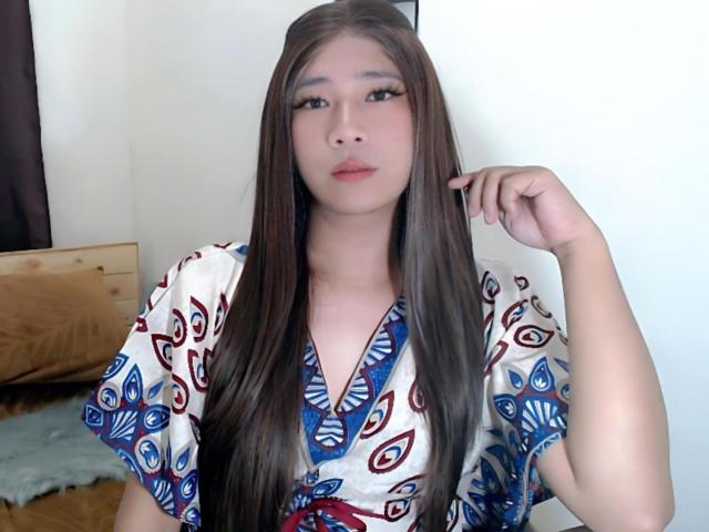 SophiaNicolex with long brown hair, wearing a white, blue, and red patterned top, looking at the camera, hand near her face, medium-sized tits, soft expression, indoor setting.