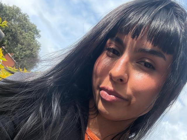 AdaWalton's face with black, straight hair and bangs, brown eyes, pink lips, and a small nose ring, looking slightly to the left with a subtle, confident smile.