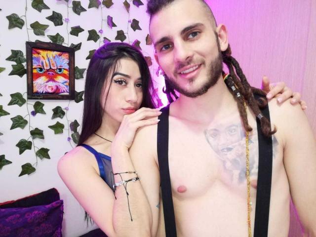 SensuaLovee with long black hair, blue tank top, holding shirtless SensuaLovee with dreadlocks, tattoo on chest, black suspenders, purple lighting, and green leaf decor.