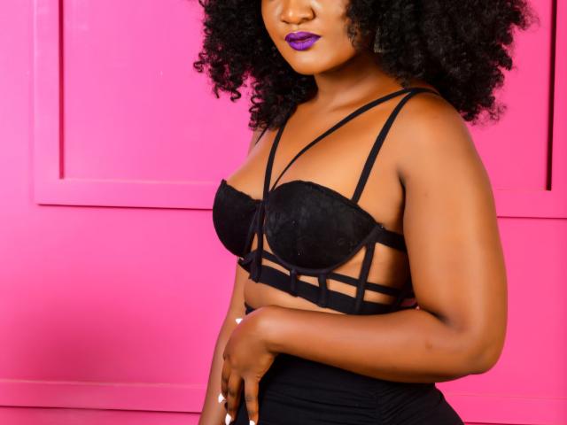 Weenie with dark brown skin, curly black hair, purple lipstick, wearing black lace bra with strappy design, showing her tits, standing against pink wall, looking confident.
