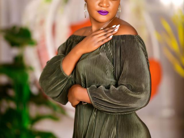 Weenie with dark skin, purple lipstick, and green off-shoulder dress, touching chest, hand with white nails, earrings, and green dress, standing, slightly turned, holding arm.
