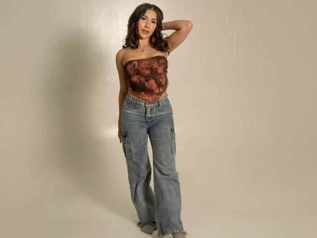 AnyaJay with brown wavy hair, wearing a strapless brown patterned top, and blue baggy jeans, standing with one hand behind her head, showing her tits and ass.