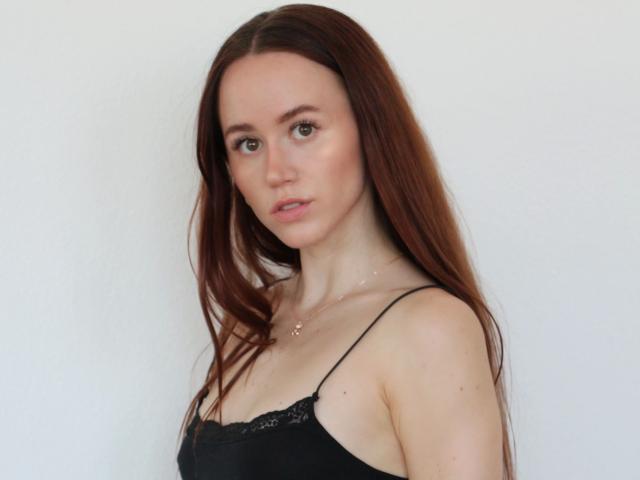 LovinLou-hot has long red hair, pale skin, and small tits, wearing a black lace-trimmed tank top, with a neutral expression and subtle makeup, against a plain white background.