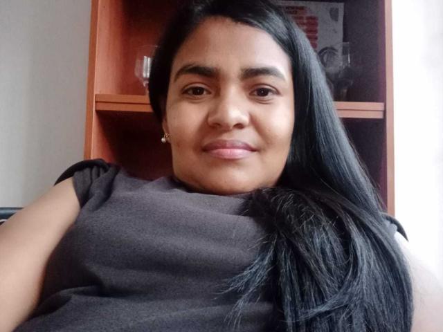 CataleyaGomez with long black hair, brown eyes, and a slight smile, wearing a black sleeveless top, showing her ass in the background. Wooden shelf with glass and book.