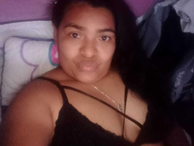 CataleyaGomez with medium brown skin and long black hair, pouting her glossy pink lips, wearing a black lace top, showing cleavage, lying in bed, looking at the camera.