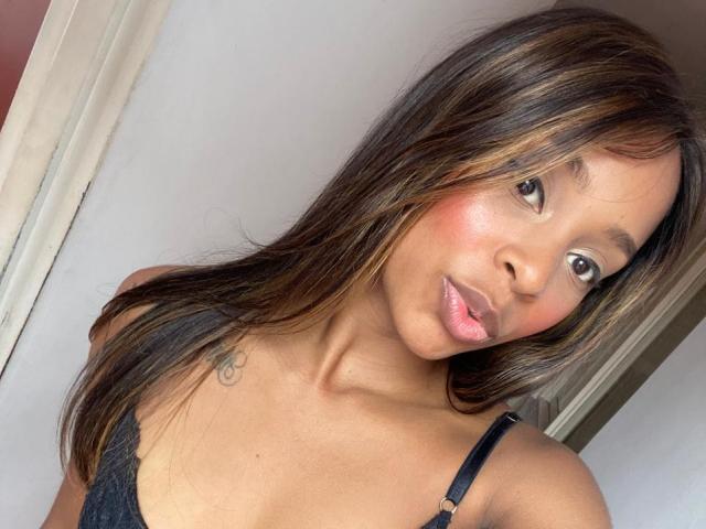 Emma, de origen latino, con cabello castaño largo, labios rosados y un tatuaje pequeño en el hombro izquierdo, vestida con un sujetador de encaje negro, sonriendo ligeramente, mostrando sus pechos y glúteos en la foto..