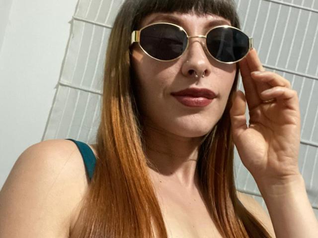 AmyLeeHot with long brown hair, nose ring, and gold aviator sunglasses, touching her ear, wearing a green top, with a slight smile, showing her lips.