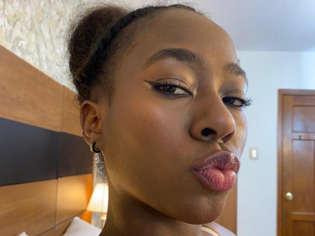 VioletGraice has dark brown skin, curly hair in a bun, thick black eyebrows, winged eyeliner, long lashes, and puffed pink lips. She wears small black earrings in a wood-paneled room.