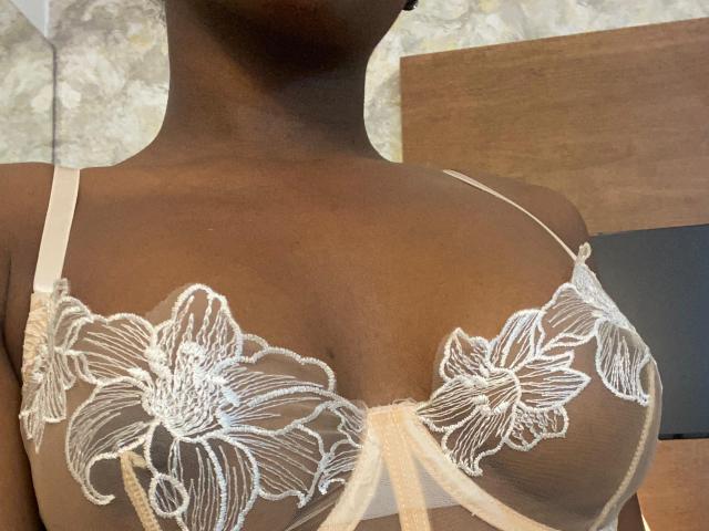 VioletGraice's dark brown skin shows off her big, round boobs in a sheer, white floral lace bra with thin straps and a small, beige bow in the center.