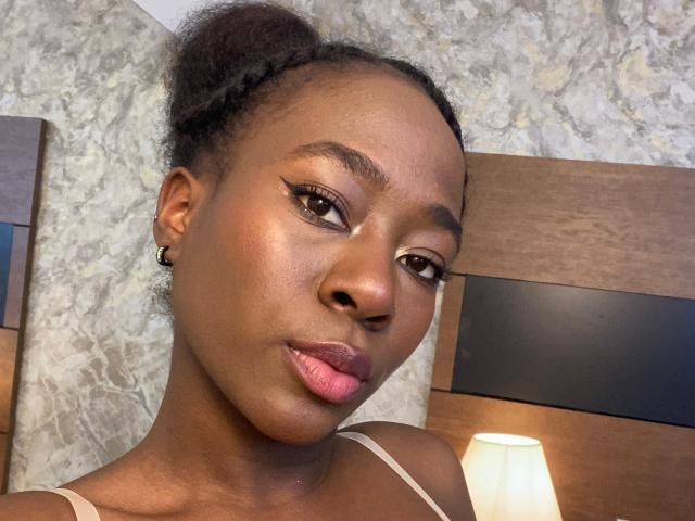 VioletGraice with dark skin, small nose, pink lips, subtle makeup, braided hair, and a serious expression, wearing a white top, in a well-lit room.