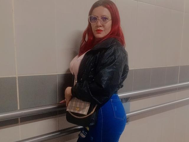 Marianiss with red hair, glasses, black leather jacket, white top, blue jeans, plaid purse, leaning on metal rail, showing her ass, standing in tiled wall hallway.
