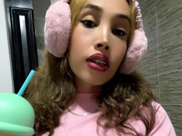 PamelaBigCoCk with pink fluffy headphones, long brown wavy hair, pink lipstick, wearing a pink shirt, holding a green cup with a blue straw, looking slightly to the side.