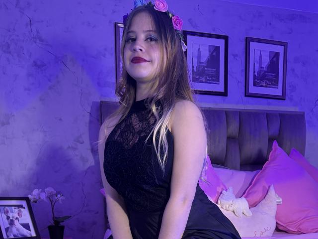 MelaniBecker with pink rose headband, black lace dress, sitting on bed, pink pillows, white stuffed toy, purple room lighting, smiling, dark red lipstick, long brown hair, light skin.