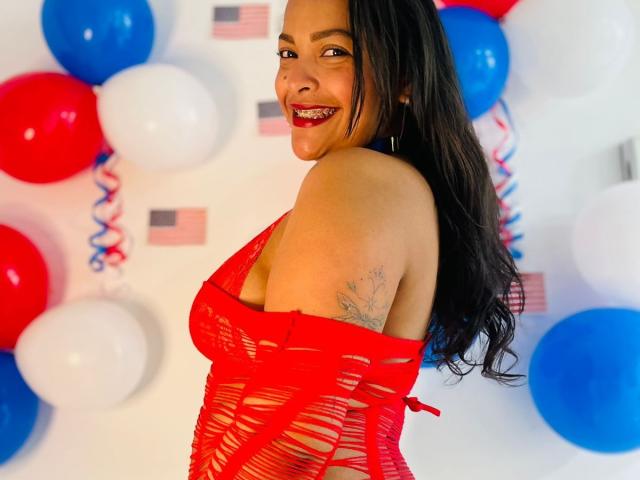 DannaRamiirez with long black hair, smiling, wearing a red, cut-out, lace-up top, showing her tits and a tattoo on her right shoulder, surrounded by red, blue, and white balloons.