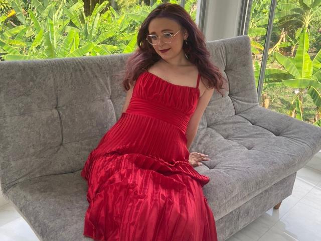 AlanisSweet with red dress, sitting on gray couch, wearing glasses, smiling, showing her tits, and her ass, with her red skirt spread out, in a bright room.