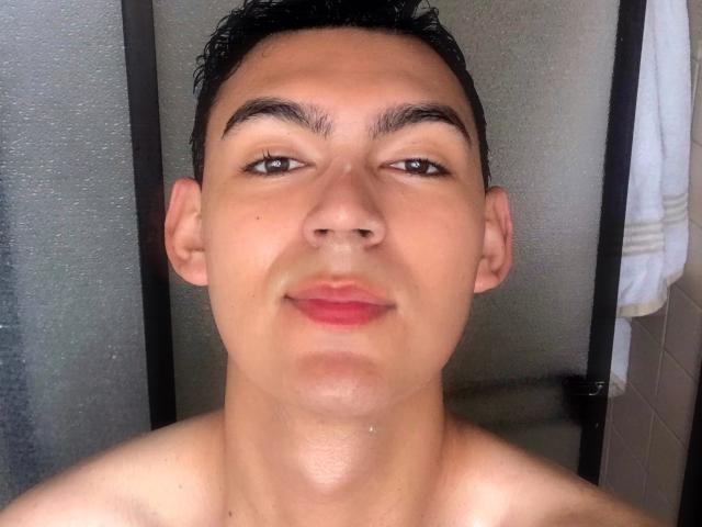 DeusBeauty, shirtless, with short black hair, brown eyes, and slightly parted pink lips, looking directly at the camera, standing in front of a frosted glass door.