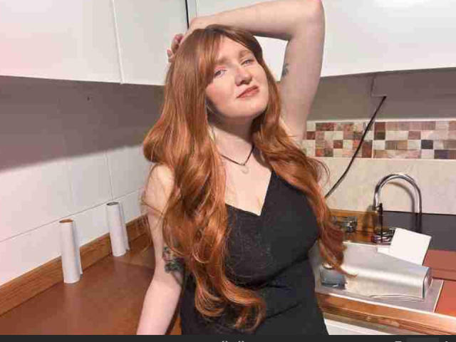 SilverLady with long red hair, black dress, arm raised, tattoo on arm, small breasts, kitchen, white cabinets, wooden countertop, sink, roll of paper towel, hand on head.