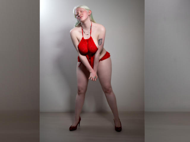 SilverLady with pale skin, blonde hair, red halter top, red bottoms, red heels, tattoo on left arm, leaning forward, hands between legs, looking down, in studio setting.