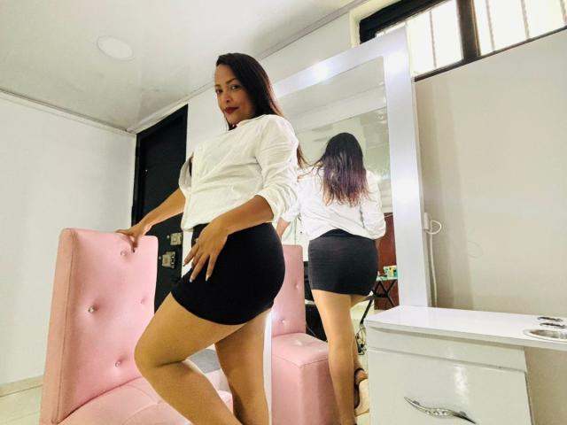 DannaRamiirez stands in a room, wearing a white shirt and black skirt, showing her ass in a mirror reflection, with a pink chair and white dresser nearby.