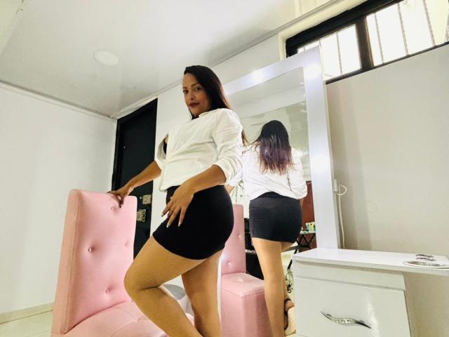 DannaRamiirez with long dark hair, white blouse, and black booty shorts, standing next to pink chair, reflection in mirror showing her ass. Room has white walls and window.
