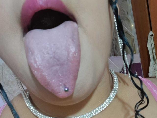 MelannieCarter's mouth is open with her tongue out, showing a pierced tongue and pink lipstick. She's wearing a diamond necklace and has blue braids.