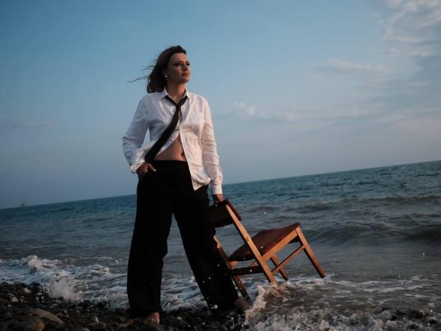 KrissSexi stands on a rocky shore with a wooden chair, wearing a white shirt, black pants, and a black tie, wind blowing through their brown hair, ocean behind.