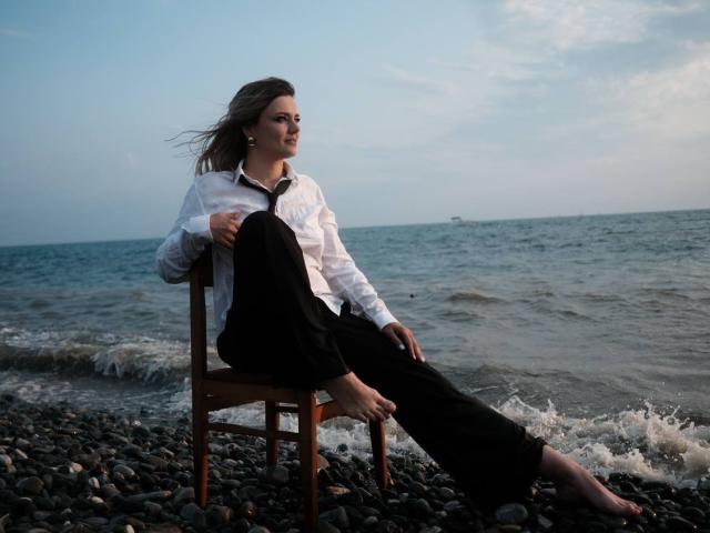 KrissSexi sits on a wooden chair by the ocean, wearing a white shirt and black pants, with her ass resting on the seat, feet in the water.