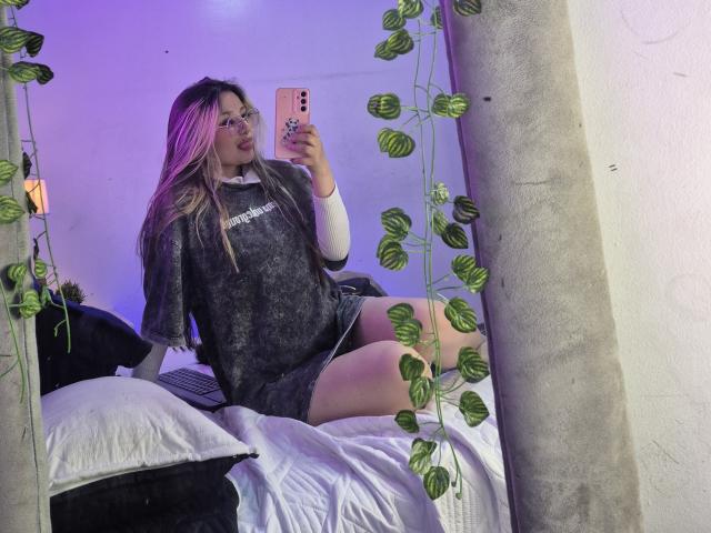 AliceStrongBeauty with pink highlights, glasses, gray dress, and white long-sleeve, sitting on bed, taking mirror selfie, green plant leaves in frame, pink phone, purple light.