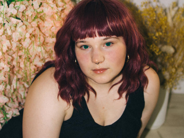 SophieLenore with red wavy hair, green eyes, nose ring, and black sleeveless top, looking at the camera with a slight pout, showing her small tits.