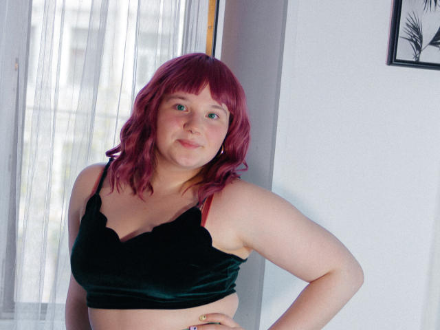 SophieLenore with pinkish-red hair, fair skin, and a cute face, wearing a black velvet bralette, standing with one hand on her hip, looking slightly shy.
