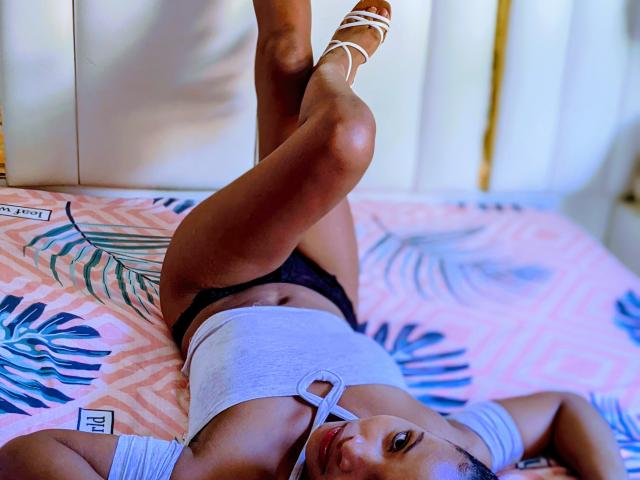 Ruckaya's legs are up, wearing white strappy sandals, black lace panties, and a white crop top with a keyhole. She's on a pink, patterned bed. Her tits are small.