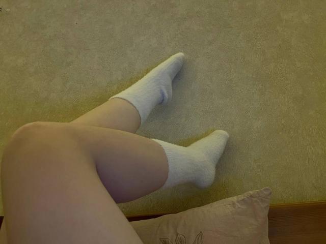 Valissias blasse Beine sind auf einem Teppich übereinandergeschlagen, sie trägt weiße Socken. Ihre nackten Oberschenkel sind sichtbar, und unten rechts befindet sich ein beiges Kissen..