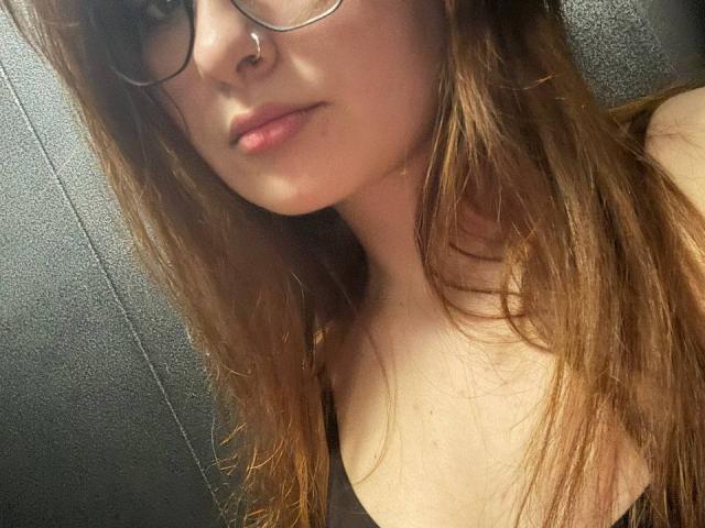 BeleMargarita with glasses, nose ring, and long brown hair, showing her tits in a dark tank top, looking at the camera with a slight pout.