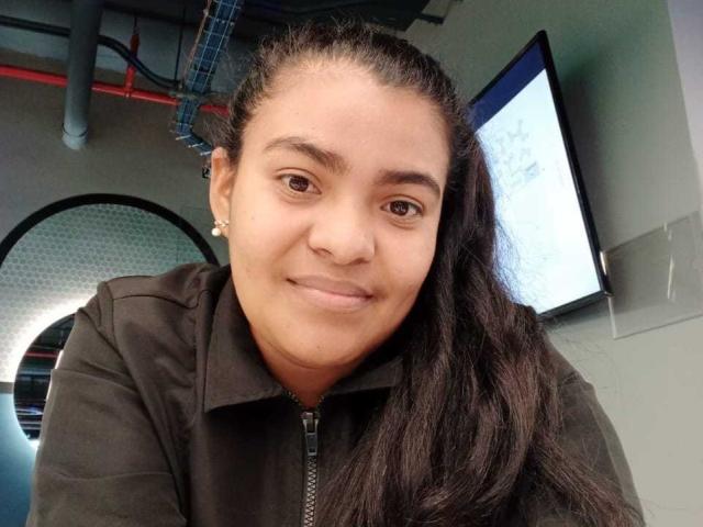 CataleyaGomez has brown eyes, long black hair, and wears a black zip-up jacket. She's smiling with a cute, small nose and light skin. A flat-screen TV is in the background.