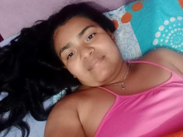 CataleyaGomez has medium brown skin, black wavy hair, and is wearing a pink spaghetti-strap top, lying on a colorful pillow, smiling slightly. Her pussy isn't visible.