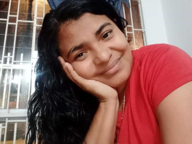CataleyaGomez with black wavy hair, red shirt, smiling softly, resting chin on hand, brown eyes, light brown skin, subtle makeup, cute and relaxed.