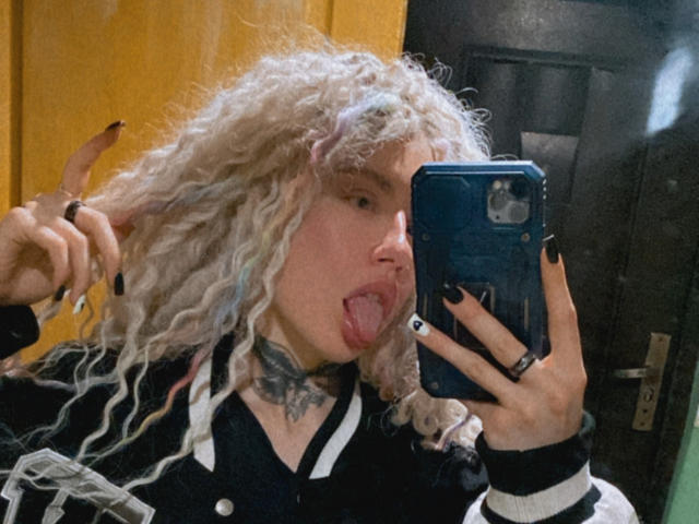 NanceDenise with curly white hair, sticking out tongue, black nails, wearing a black and white jacket, holding a blue phone, and showing off a tattooed neck.