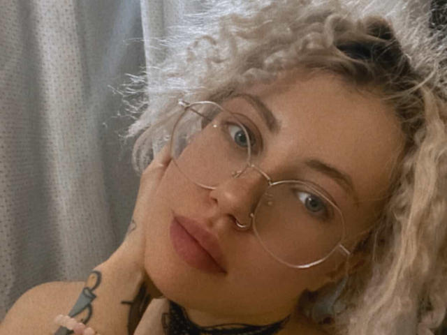 NanceDenise has curly blonde hair, wears round glasses, and has a nose ring. She's touching her cheek with a tattoo visible on her neck. Soft lighting.
