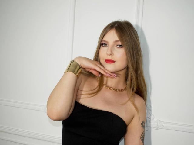 SaraMayery with long brown hair, red lipstick, black strapless top, gold bracelet, gold necklace, and tattoo on right arm, touching chin, smiling.