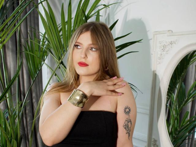 SaraMayery with light brown hair, red lipstick, black strapless top, gold bracelet, and tattoo on right arm, standing near green plants, looking to the side with a calm expression.
