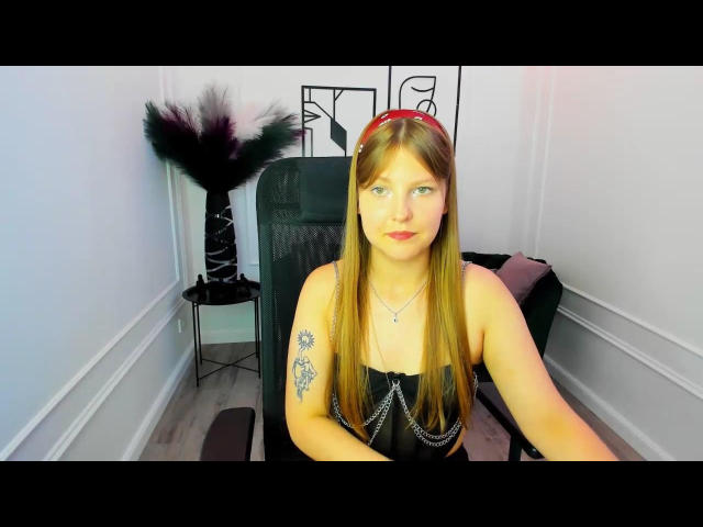 SaraMayery, with long blonde hair, wears a black strapless dress, red headband, and necklace, showing tattoos on her right arm, sitting in a black chair.