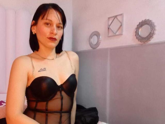 ViolettaRosse with pale skin, black bob, dark lipstick, wearing black sheer lingerie with strap chains, small tits, tattoo on chest, standing in a room, looking at camera.