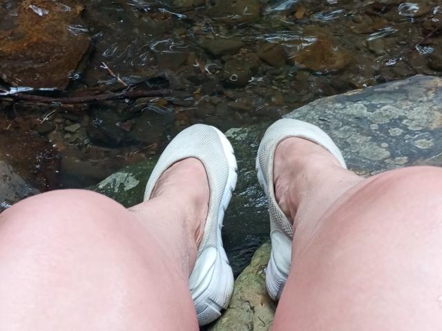 KelliRedhaer's white sneakers and pale, slightly hairy legs are in the water, with rocks and clear water visible below. Her feet are slightly apart, showing her toes.