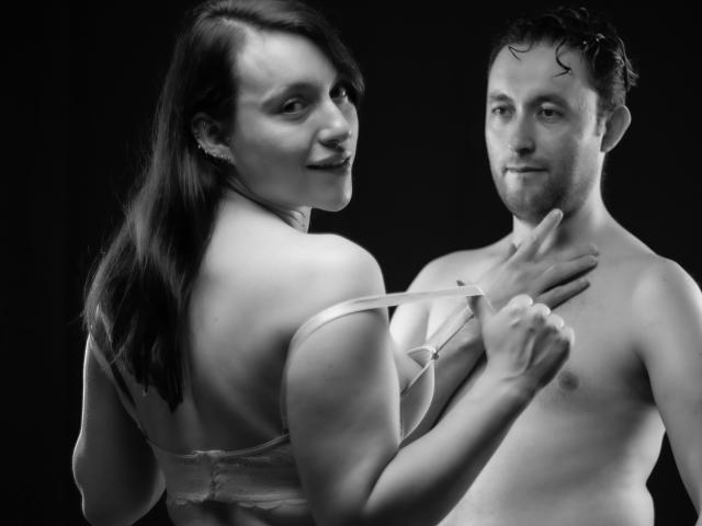 JeremmyAndStacy, una mujer con los senos al descubierto en un sujetador, tirando de una correa de su espalda, sonriendo a un Jeremmy sin camisa con un dedo en su barbilla, en blanco y negro..