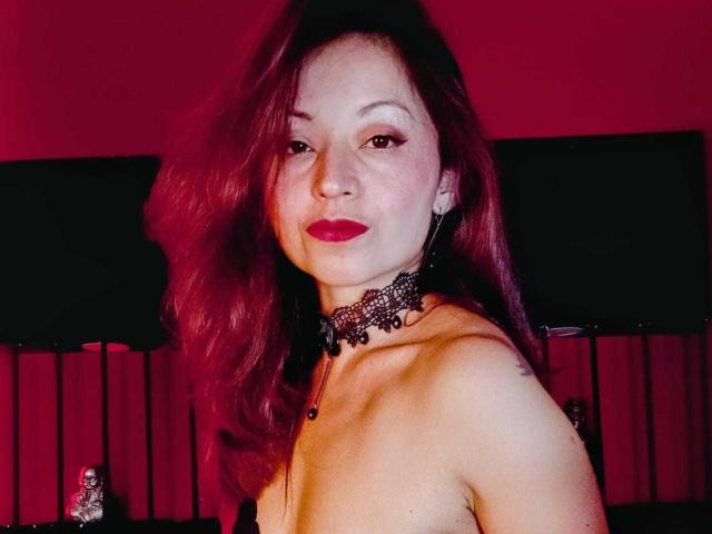 AngelinaBlanche with red hair, black lace choker, and red lipstick, topless, showing her tits, slight smile, tattoo on right shoulder, red background.