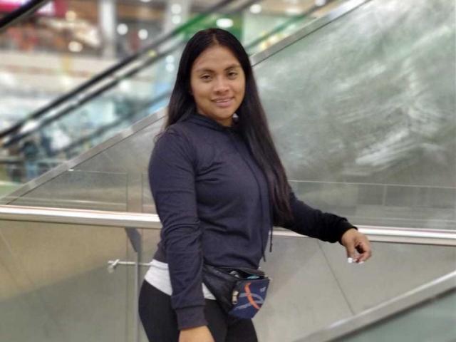 DahianaColeman with long black hair, wearing a navy hoodie, white shirt, black pants, and a blue bag, leaning against a glass railing, smiling. Her ass is slightly visible.