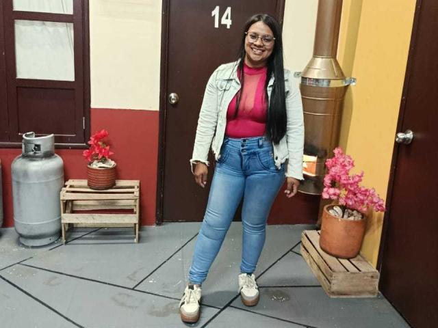 DahianaColeman with long black hair, glasses, red turtleneck, white jacket, blue jeans, and white platform shoes, smiling, standing in a room with potted plants and a metal tank.